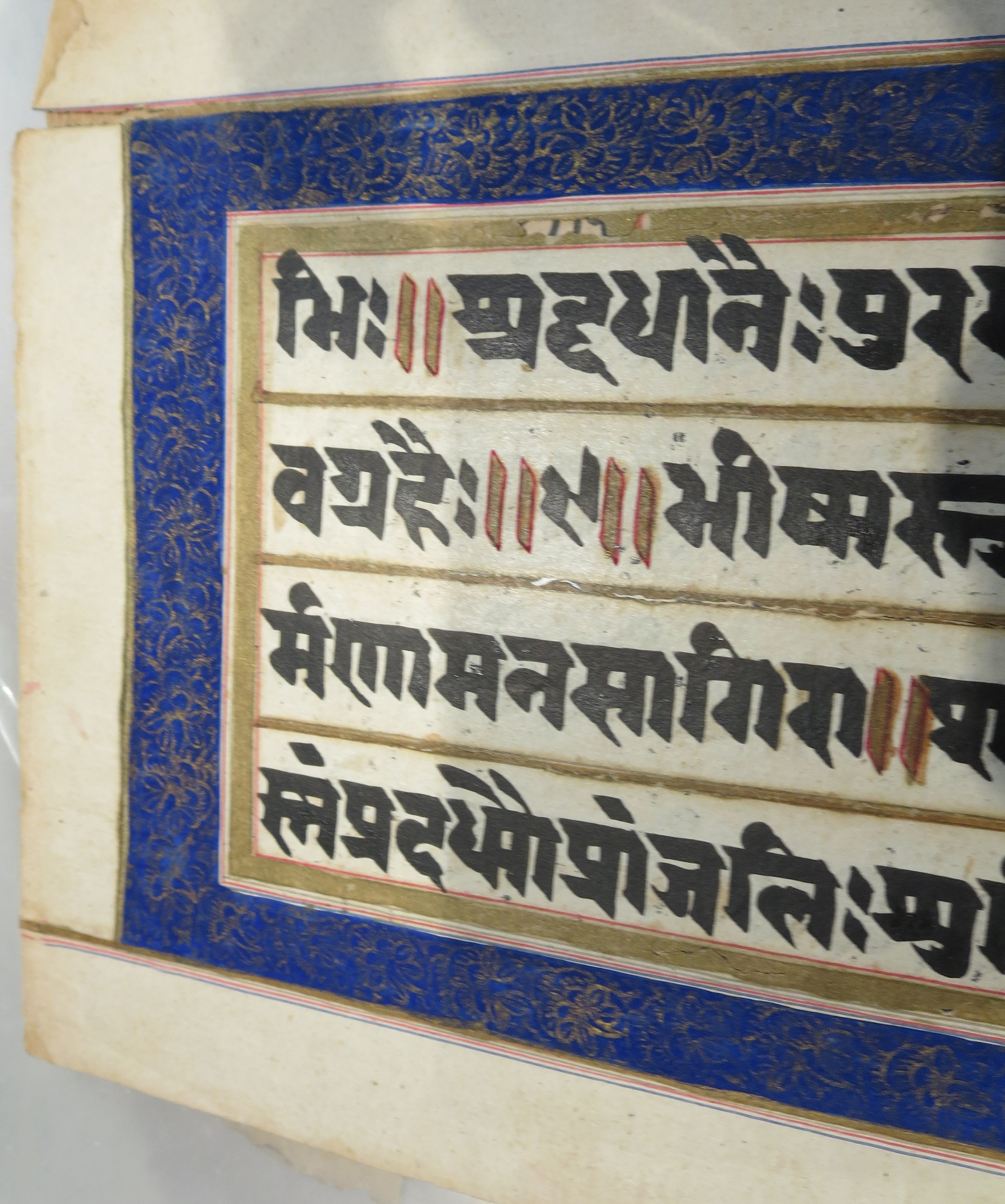 Blue-Bordered Side of the Paper Leaf in Sanskrit, seen from an angle within the transparent archival polypropylene slip-in protector. Image cropped to left-hand side of the column of text, plus outer margin.Private Collection. Photograph by Mildred Budny.